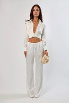 Woman in white satin set: cropped wrap top and wide-leg drawstring pants with a neutral woven handbag.